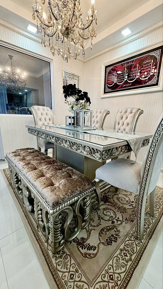 A luxurious dining room set featuring a French style mahogany wood dining table with ornate detailing, accompanied by four French style dining chairs and a scroll bench, all carpeted in a lavish room with a chandelier and intricate wall art.
