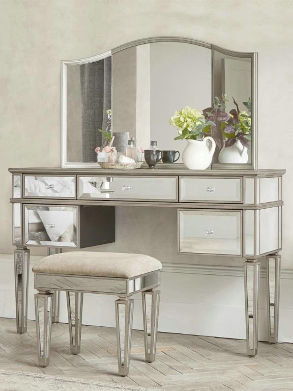 A luxurious vanity set with a mirrored surface, including a table with drawers, a folding mirror, and a stool. The set has a silver and white color scheme.