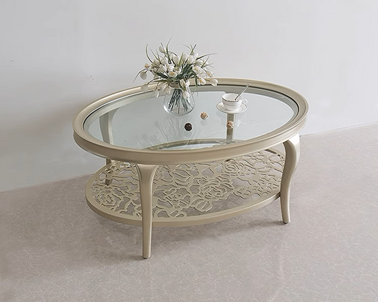 A round, modern Victorian style coffee table with a glass top and carved wooden legs, displayed on a white floor with a vase of flowers on top and a candle holder beside it.