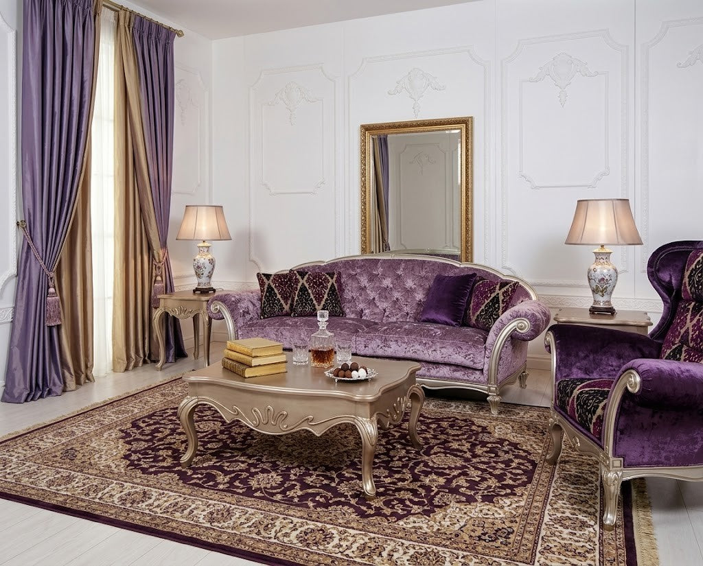 An elegant living room featuring a tufted purple velvet sofa, a matching purple armchair, a carved silver coffee table, and a large patterned oriental rug on a white paneled room.
