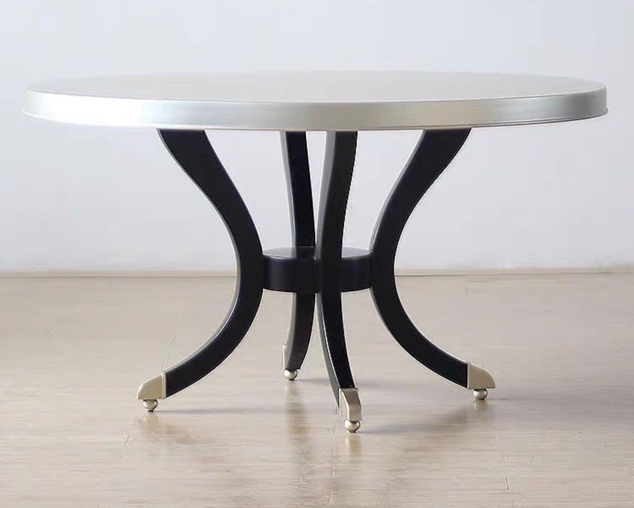 A modern luxury style round dining table with a silver tabletop and black wooden frame with gold accents, presented by Dainté Living.