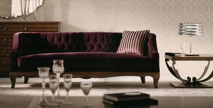 A tufted velvet sofa in deep burgundy with wooden legs, styled in a modern living room setting with a striped throw pillow, side table, and glassware.