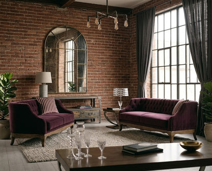 A styled living room featuring two deep purple velvet sofas with wooden legs, set against a rustic brick wall with a large arched mirror and tall multi-pane windows with dark gray curtains.