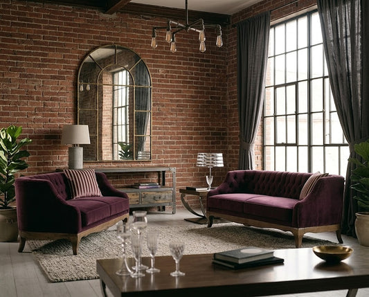 A styled living room featuring two deep purple velvet sofas with wooden legs, set against a rustic brick wall with a large arched mirror and tall multi-pane windows with dark gray curtains.