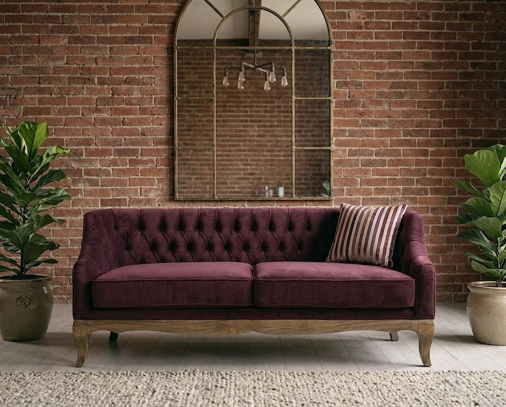 A tufted two-seat sofa upholstered in deep burgundy velvet with a light natural wood frame, set against a brick wall with a large arched mirror and potted plants.