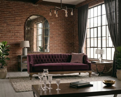 A deep burgundy tufted velvet sofa with wooden legs in a rustic living room with exposed brick walls, a large arched mirror, and tall windows with dark curtains.