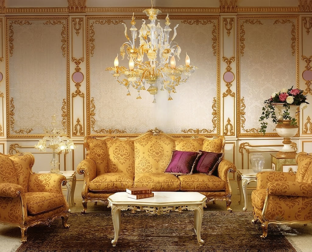 An ornate luxury living room set featuring a gold damask sofa, two matching armchairs, a white marble-top coffee table with gold legs, and a large crystal chandelier against a decorated gold and beige paneled wall.