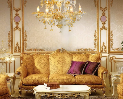 An ornate gold and beige damask sofa in a luxurious room decorated with gold molding, a large crystal chandelier, and purple accent pillows.
