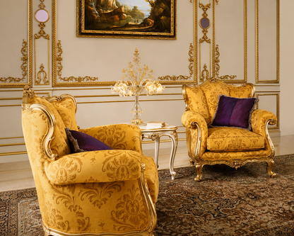 Two ornate yellow damask armchairs with gold-trimmed cabriole legs, arranged in a luxurious room with a small silver side table, a crystal chandelier, and a patterned rug.
