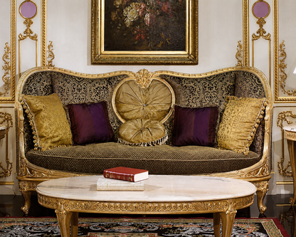An ornate baroque-style sofa with a gilded frame, patterned upholstery, and decorative pillows, set in a luxurious room with a matching carved coffee table and framed wall art.