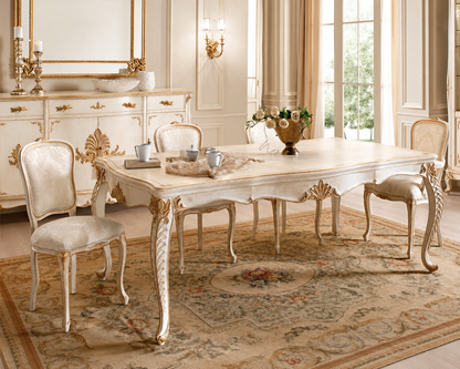 An elegant French-style dining room set featuring a rectangular table and four matching chairs in a cream and gold finish, arranged on a patterned area rug in a luxurious room with classical wall moldings and windows.