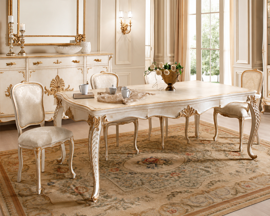 An elegant French-style dining room set featuring a rectangular table and four matching chairs in a cream and gold finish, arranged on a patterned area rug in a luxurious room with classical wall moldings and windows.