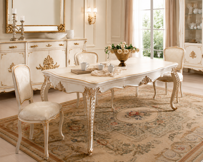 An elegant white carved dining table with four matching upholstered chairs, set on a large patterned area rug in a luxurious cream-colored dining room with gold accents.