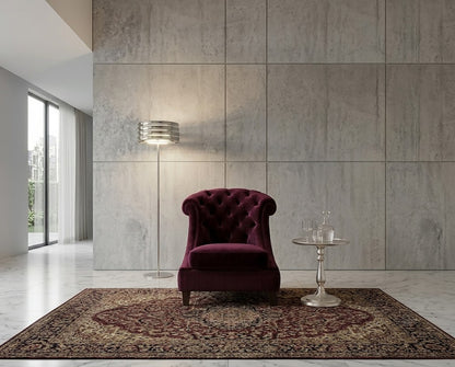 A deep burgundy velvet tufted armchair is placed in a modern interior setting on a patterned area rug, with a silver floor lamp and a small round side table beside it against a large textured gray wall.