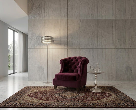 A deep burgundy tufted velvet armchair sits on a patterned area rug in a modern living room with a concrete-paneled wall, a floor lamp, and a small side table.