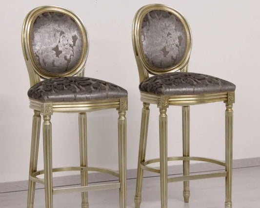 Two ornate bar stools with gold frames and gray upholstered seats on a plain background.