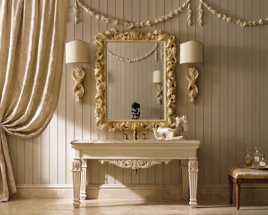 An ornate cream-colored console table with a matching decorative mirror and two matching wall sconces, set against a beige paneled wall with draped curtains.