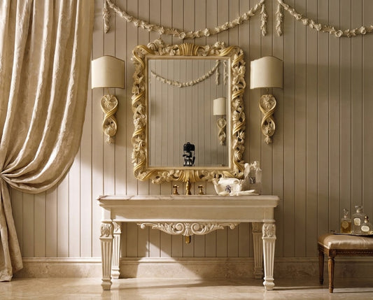 An ornate cream-colored console table with a matching decorative mirror and two matching wall sconces, set against a beige paneled wall with draped curtains.