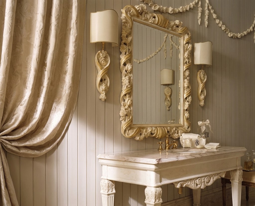 An ornate gold-framed wall mirror with a cream fabric lampshade sconce mounted on a paneled wall above a matching cream console table, flanked by two identical sconces and draped champagne-colored curtains.