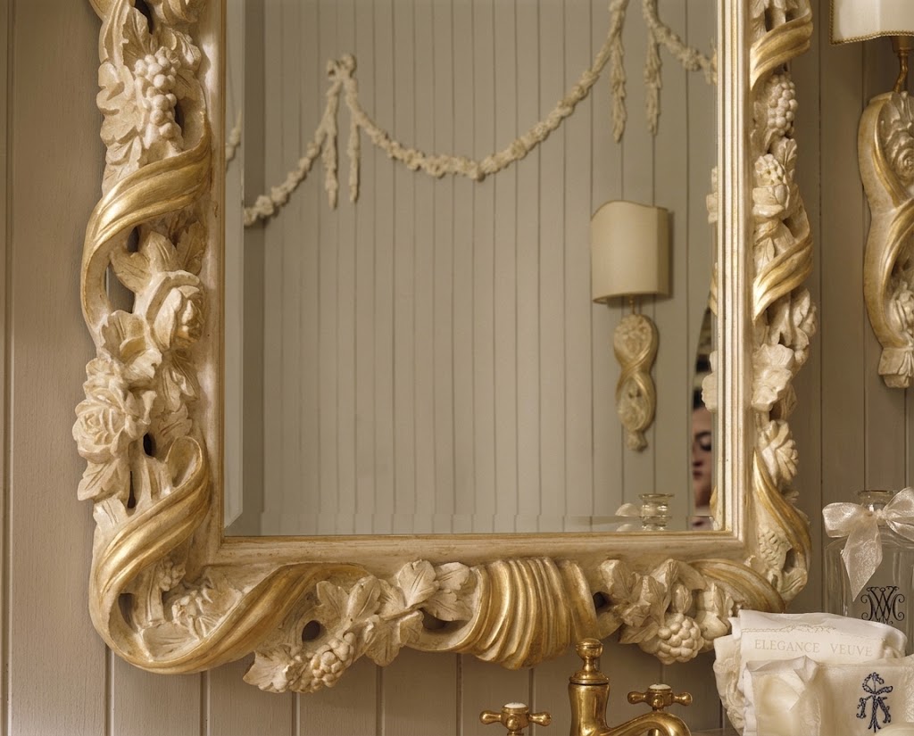 An ornate gold and beige carved decorative wall mirror with intricate floral and scrollwork details, mounted on a paneled wall above a brass faucet.