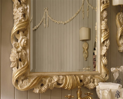An ornate gold and beige carved decorative wall mirror with intricate floral and scrollwork details, mounted on a paneled wall above a brass faucet.