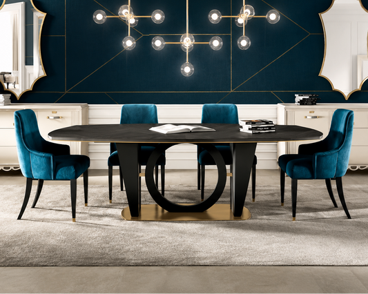 A modern dining room setup featuring a dark oval dining table with a unique circular base, surrounded by four teal velvet chairs. The room has a dark teal geometric wall with gold lines, two gold-framed mirrors, and a multi-arm brass pendant chandelier.