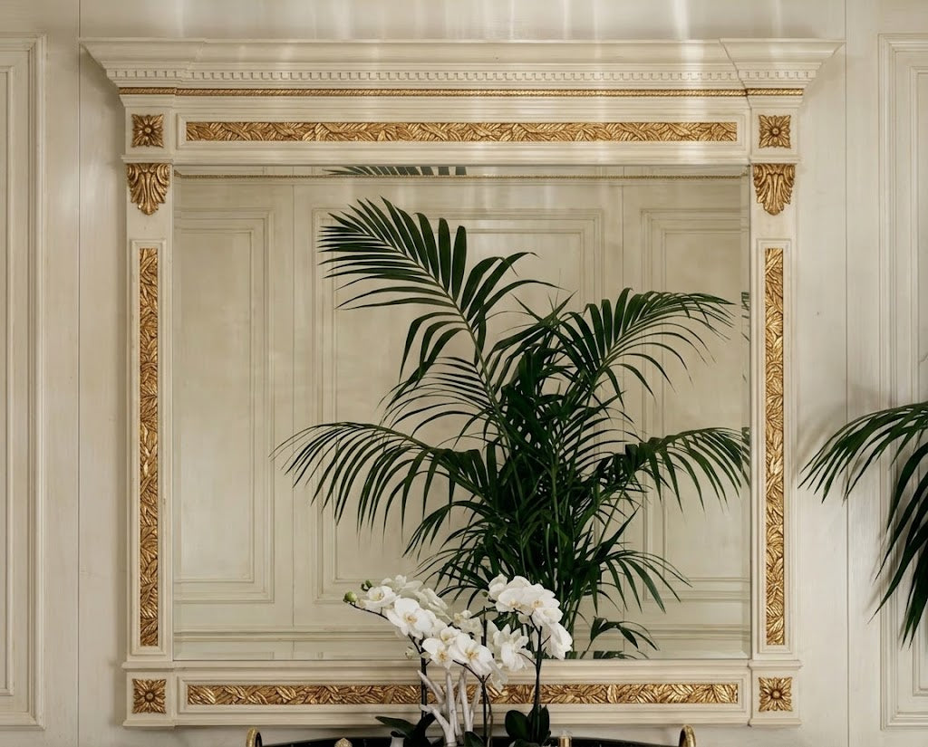 A large rectangular wall mirror with an ornate cream and gold frame, reflecting a green palm plant and white orchids in a decorated interior setting.