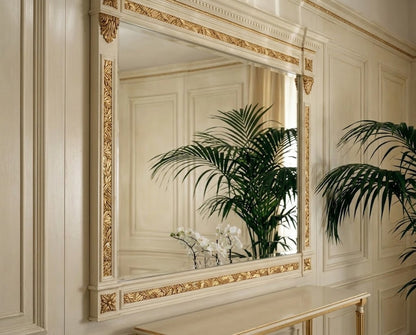 An ornate cream-colored wall mirror with intricate gold gilded detailing around the frame, mounted on a matching cream paneled wall. The reflection shows green palm leaves and white flowers, and a gold-framed console table is visible at the bottom.