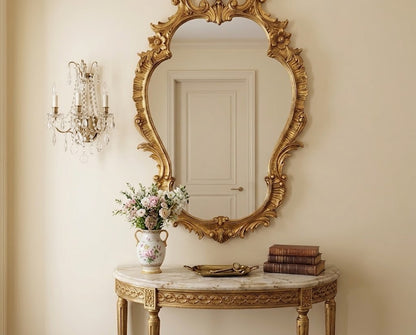 An ornate gold-framed oval wall mirror hangs above a carved console table in a cream-colored room. A floral arrangement in a vase and a stack of books sit on the table, and a crystal candlestick sconce is visible on the left wall.