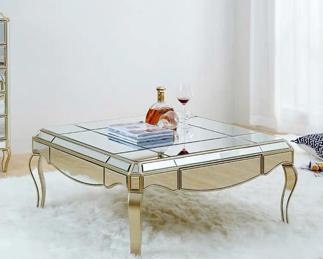 A luxurious coffee table with a mirrored surface and gold finish accents, displayed in a bright room with a white rug.