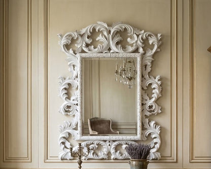 An ornate white carved mirror with a baroque-style frame hangs above a matching white three-drawer dresser in a classic room with beige paneled walls, a wooden cabinet, and a chair.