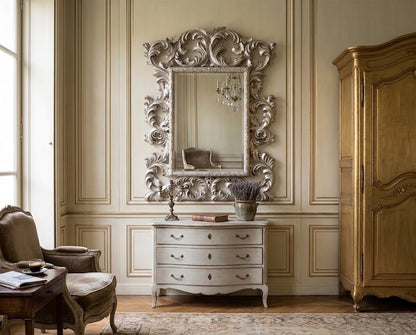 An ornate silver mirror with a highly detailed baroque-style carved frame, mounted on a beige paneled wall above a white three-drawer console table in a classic European-style room.