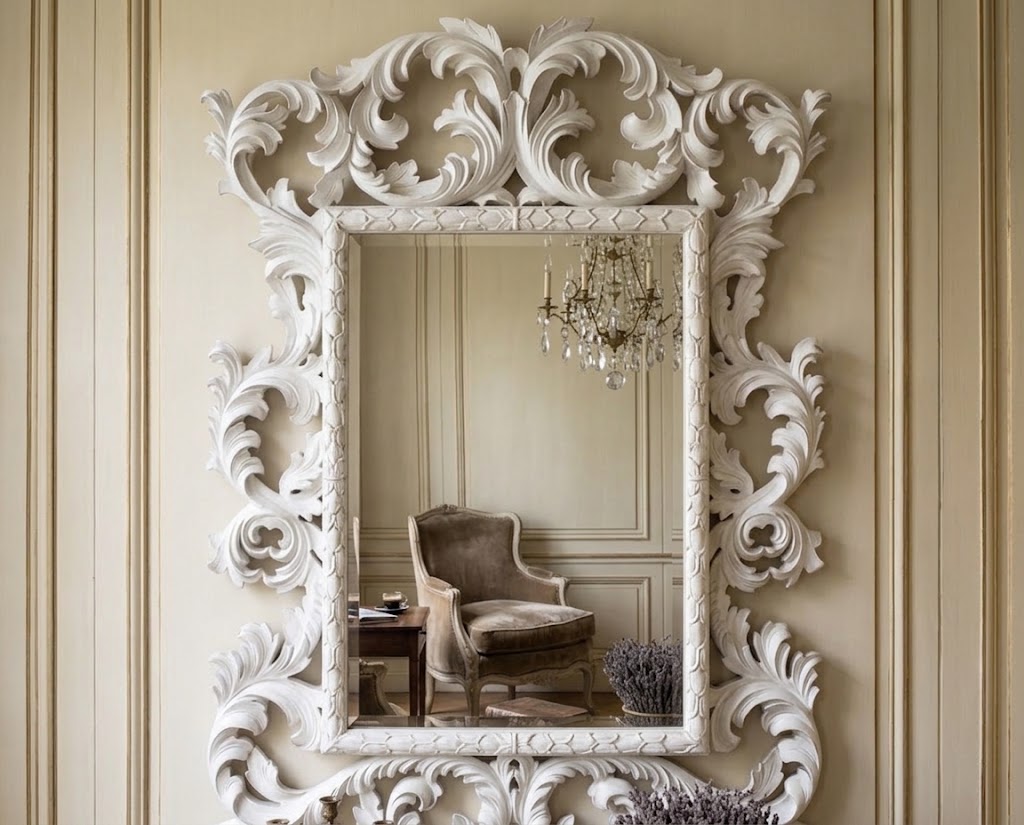 An ornate white carved wall mirror with intricate scrollwork, mounted on a beige paneled wall. The mirror reflects a classic armchair and a crystal chandelier.