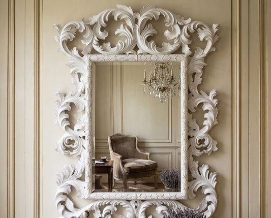 An ornate white carved wall mirror with intricate scrollwork, mounted on a beige paneled wall. The mirror reflects a classic armchair and a crystal chandelier.
