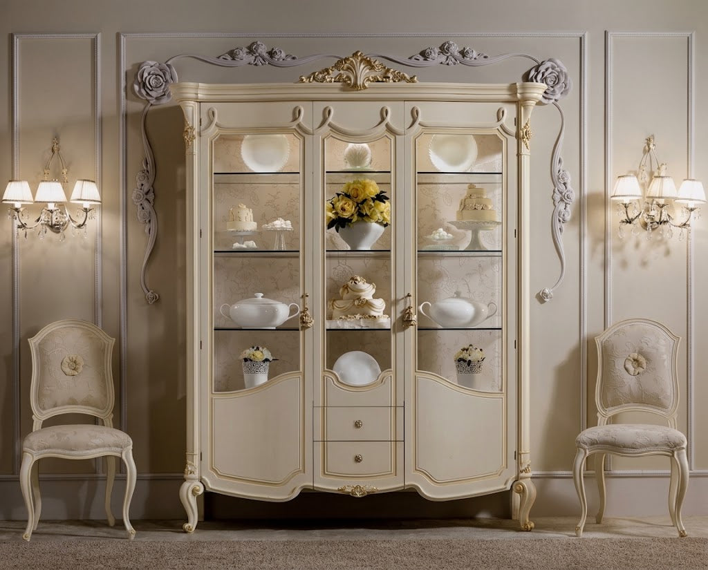 An ornate cream-colored Italian-style display cabinet with glass doors, styled in a classic elegant room with two matching chairs and wall sconces.