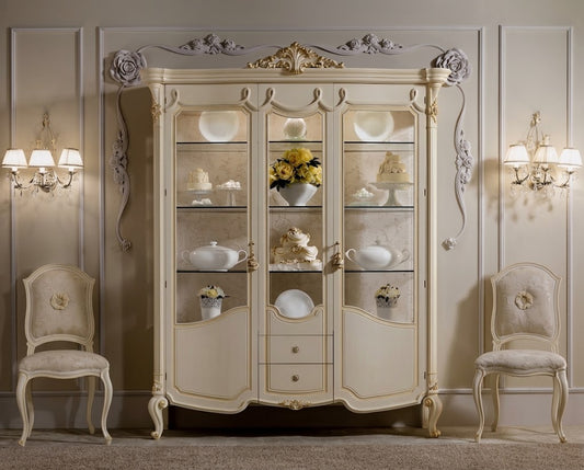 An ornate cream-colored Italian-style display cabinet with glass doors, styled in a classic elegant room with two matching chairs and wall sconces.