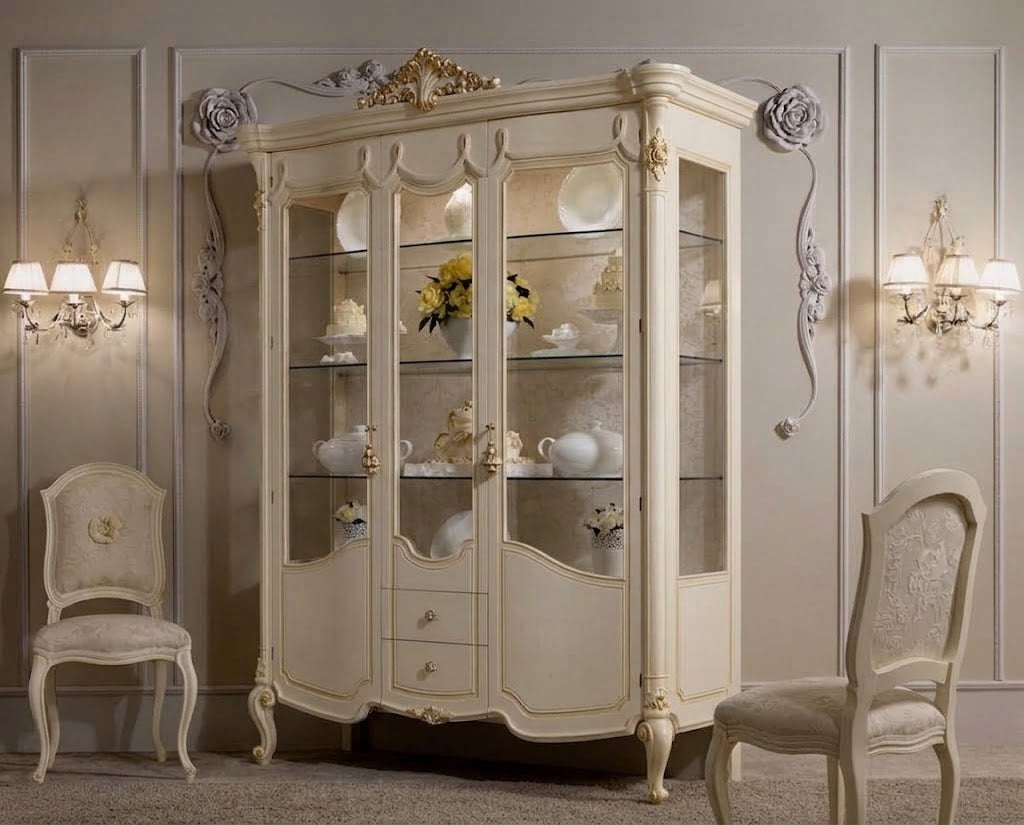 An ornate cream-colored corner display cabinet with glass doors, styled in a classic elegant room with matching chairs and wall sconces.