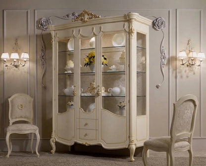 An ornate cream-colored corner display cabinet with glass doors, styled in a classic elegant room with matching chairs and wall sconces.