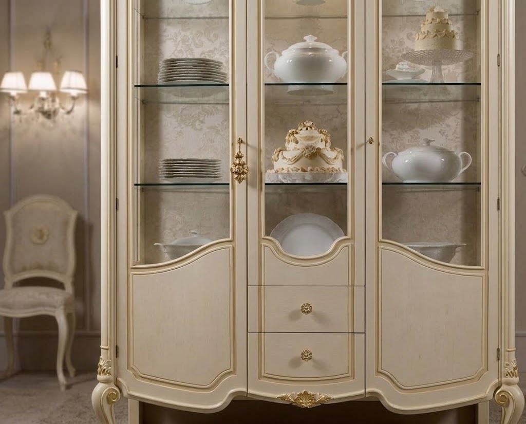 An elegant cream-colored display cabinet with gold trim and glass doors, showcasing white porcelain dishes and a decorative cake inside, set in a classic interior room.