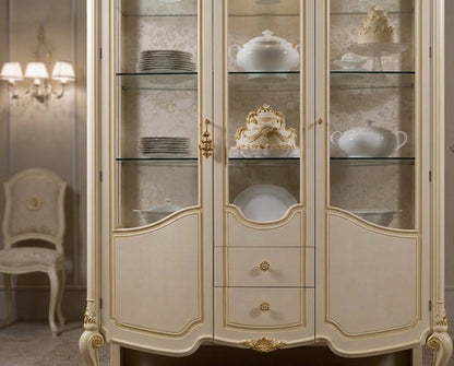 An elegant cream-colored display cabinet with gold trim and glass doors, showcasing white porcelain dishes and a decorative cake inside, set in a classic interior room.