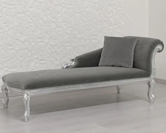 Gray velvet chaise lounge with silver legs against a textured white wall.