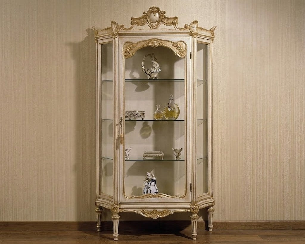 An ornate vintage-style glass display cabinet with a distressed gold and beige frame, shown against a beige textured wall and wooden floor.