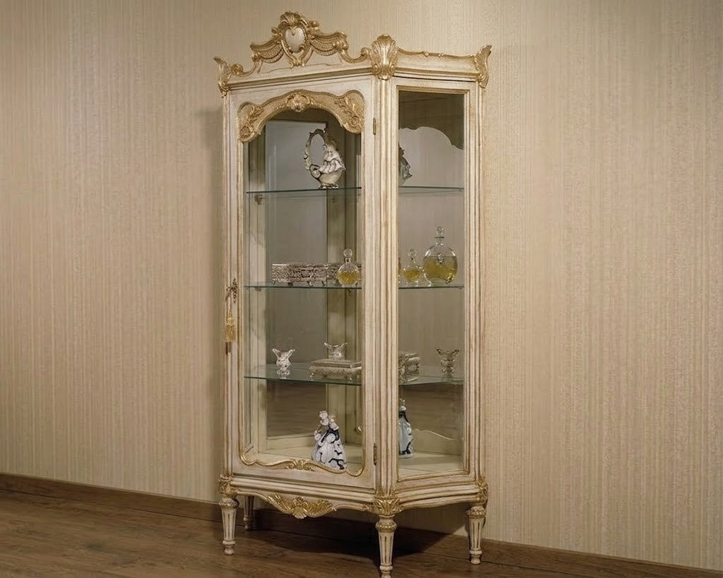 An ornate vintage-style glass display cabinet with a distressed cream and gold frame, standing against a beige textured wall and wooden floor.