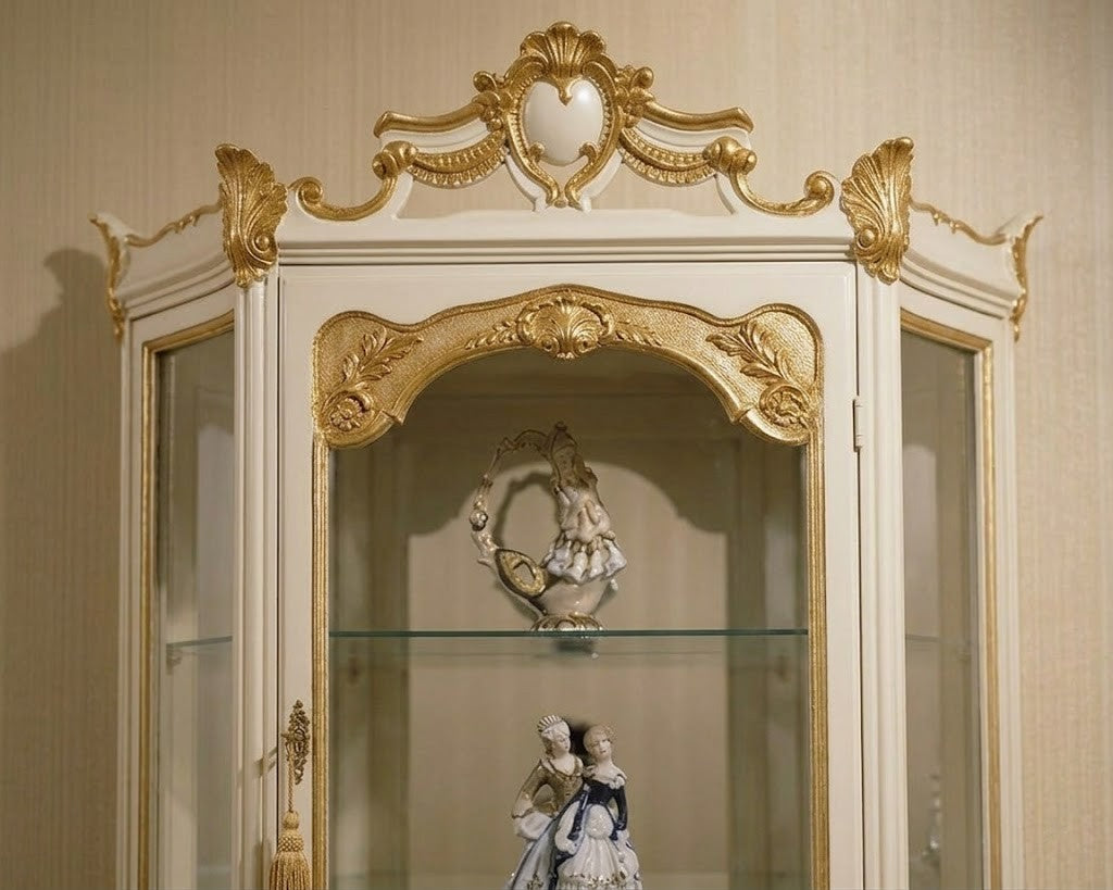 An ornate cream and gold corner display cabinet with glass doors, showcasing decorative porcelain figurines inside against a beige textured wall.