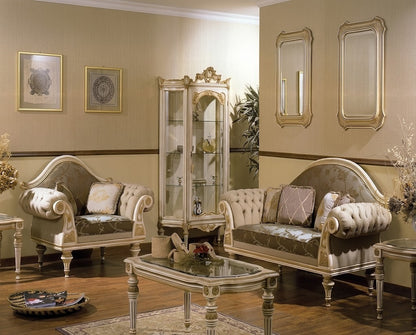 An ornate classic living room furniture set featuring a tufted chaise lounge, a matching sofa, a glass-top coffee table, and a tall decorative glass display cabinet, all in cream and gold upholstery with carved wood details.