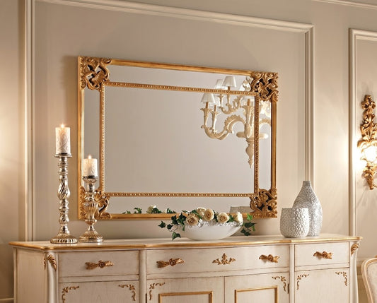 An ornate rectangular wall mirror with a gilded gold frame, mounted on a beige paneled wall above a matching cream and gold console table decorated with candles, a bowl of fruit, and a textured vase.