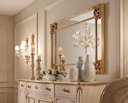 An ornate gold-framed rectangular wall mirror hanging above a cream console table with gold trim, styled with candles, vases, and flowers in an elegant interior room.