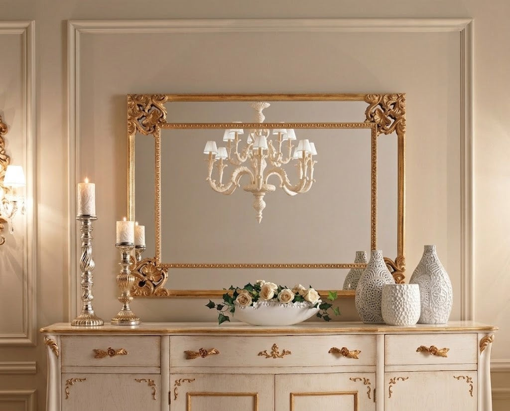 An ornate rectangular wall mirror with a gilded gold frame, mounted on a beige paneled wall above a matching cream-colored console table decorated with candles, vases, and a floral arrangement.