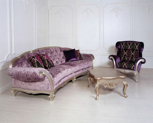 A luxurious purple velvet sofa with ornate gold-toned legs, accompanied by a matching purple and black patterned armchair and a small matching side table, set against a white paneled wall.