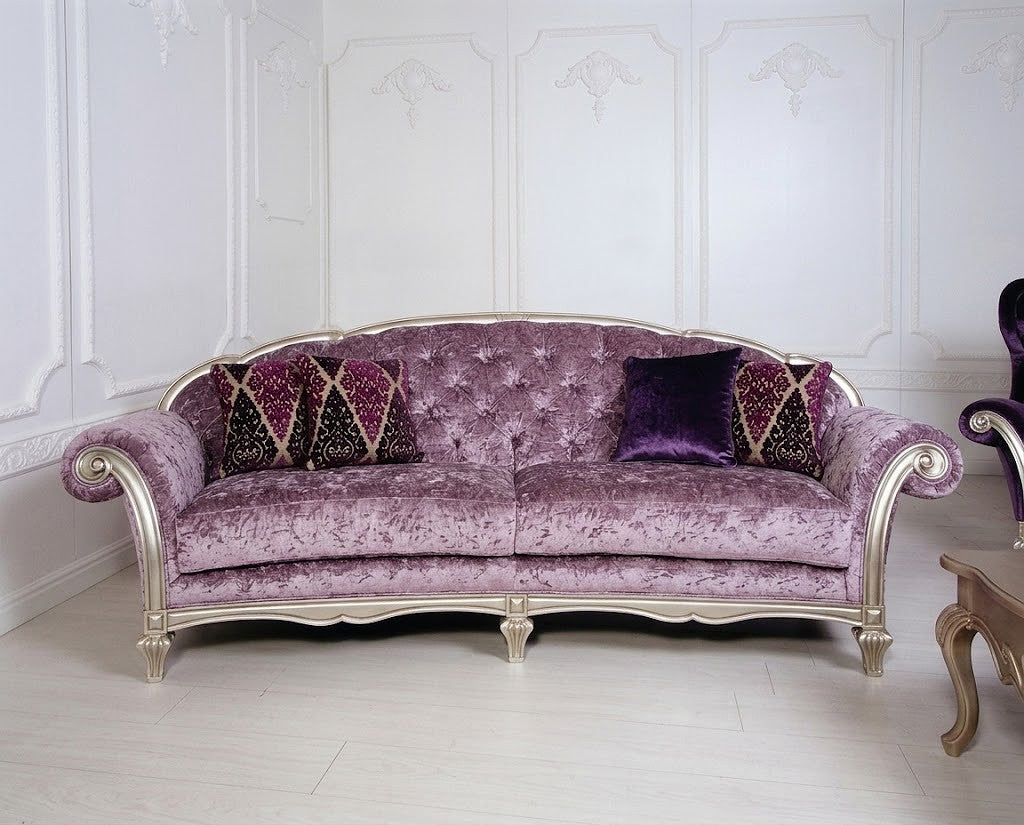 A luxurious tufted velvet sofa in lavender purple with ornate silver-gilt carved arms and legs, styled with two patterned accent pillows and one solid purple throw pillow in a white paneled room.